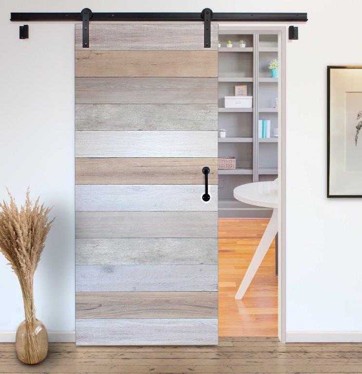 Cascade sliding barn door with a horizontal wood-plank pattern in light beige, gray, and white tones, shown installed on a black metal track in a bright room with a visible shelving unit behind it.