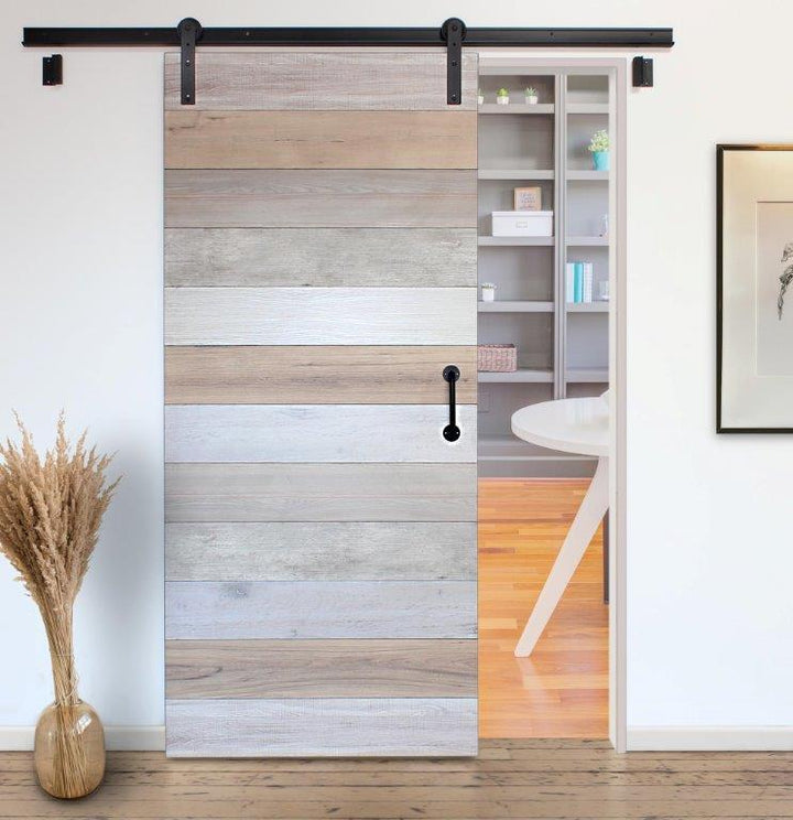 Cascade sliding barn door with a horizontal wood-plank pattern in light beige, gray, and white tones, shown installed on a black metal track in a bright room with a visible shelving unit behind it.