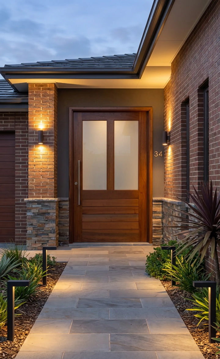 horizontal slatted Meranti pivot door installed in a brick home exterior at dusk, with frosted glass panels, warm timber stain, garden path, and wall lighting.
